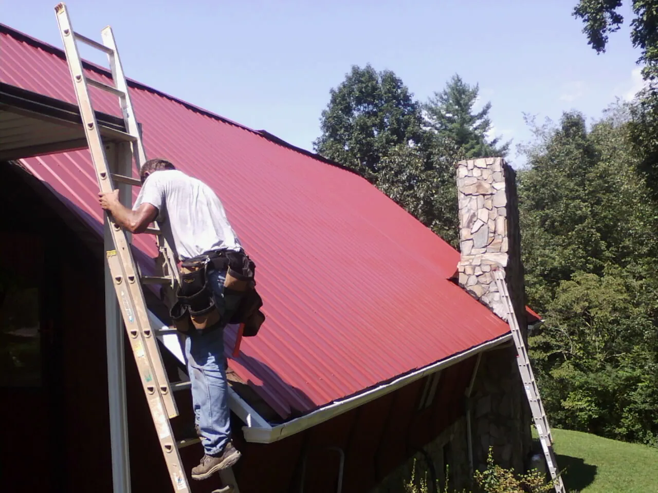 Roofer on a ladder beside a red metal roof and stone chimney — Soffit & Fascia Repair by Roofing Brothers in Point Pleasant Beach